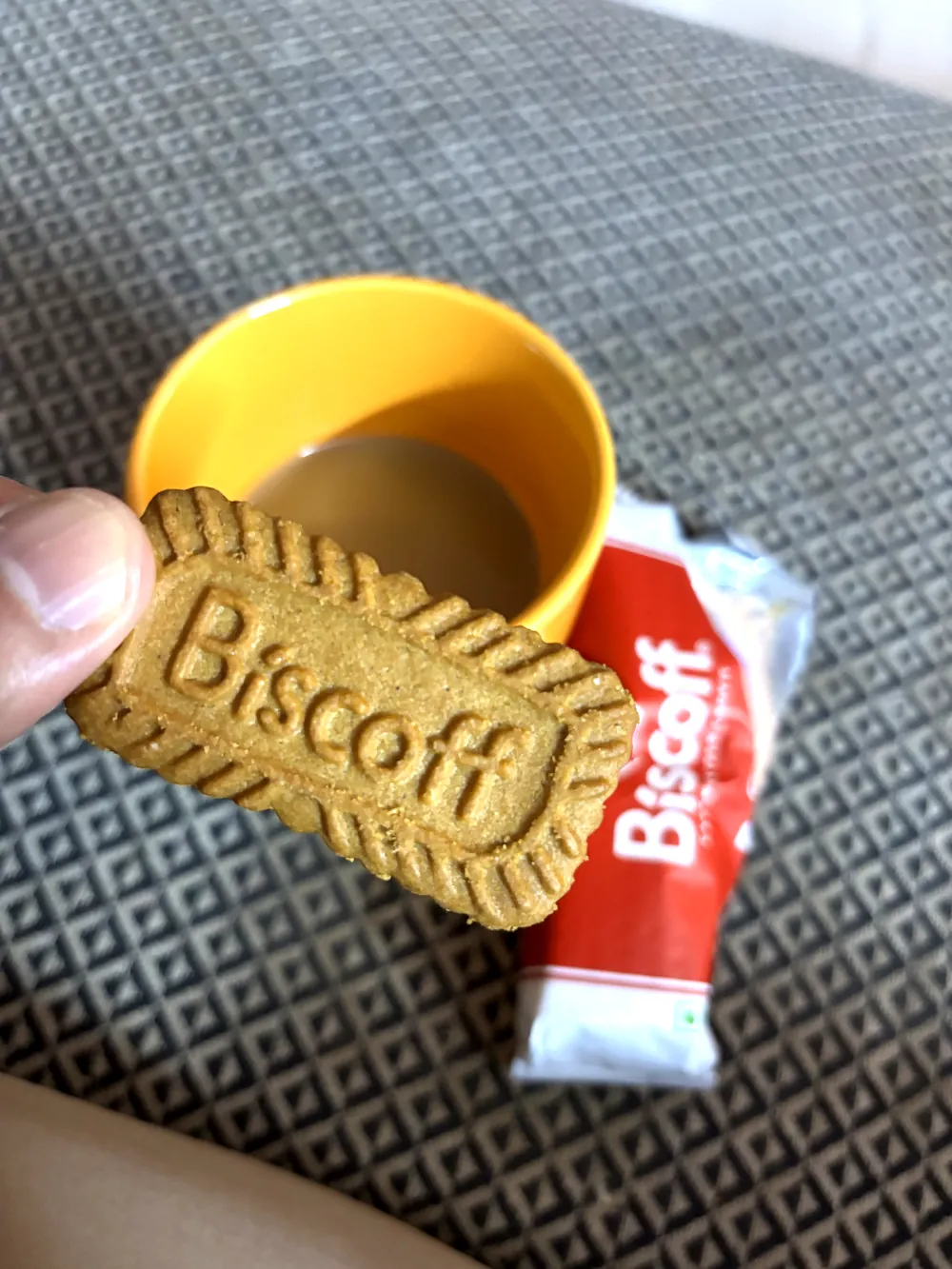 A Lotus Biscoff biscuit with its embossed logo, held beside a cup of chai