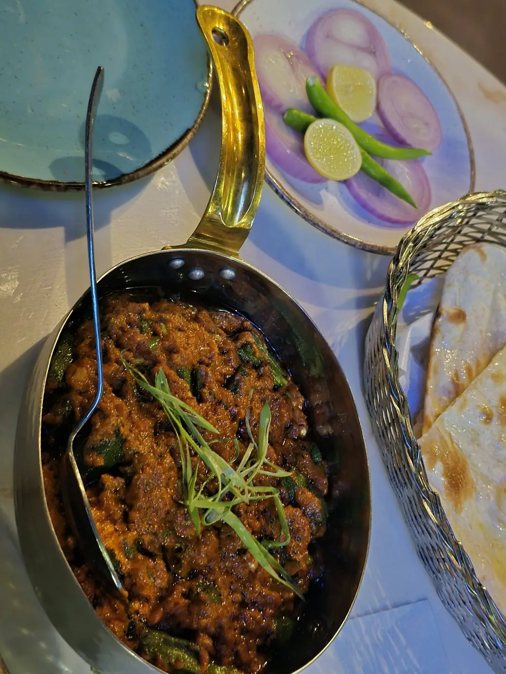 Bhindi masala served in a brass-handled pan with naan, sliced onion, lime and green chilli