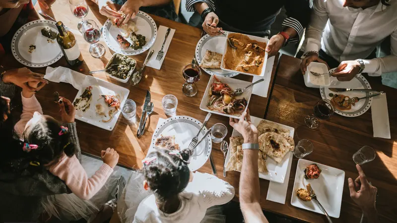 Overhead view of a shared Indian meal, with several people reaching across a wooden table of curries, naan and grilled dishes