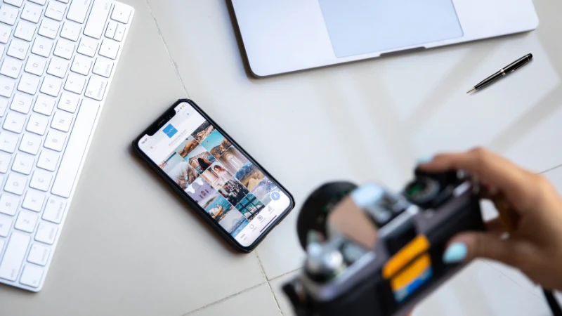 Creator holding a camera above a desk with a laptop, keyboard and a phone showing a photo grid