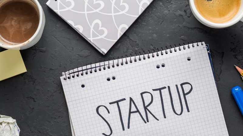 Notebook on a dark desk with the word Startup written on it, beside coffee cups, a pen and a phone